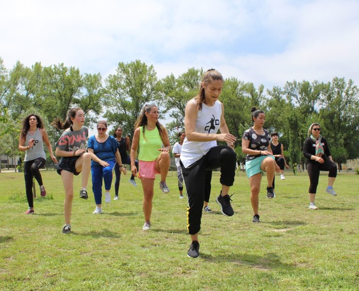 Instrutora de fitness com uma camisola branca e umas calças pretas apoiada numa só perna a coreografar uma equipa de dançarinos num jardim.